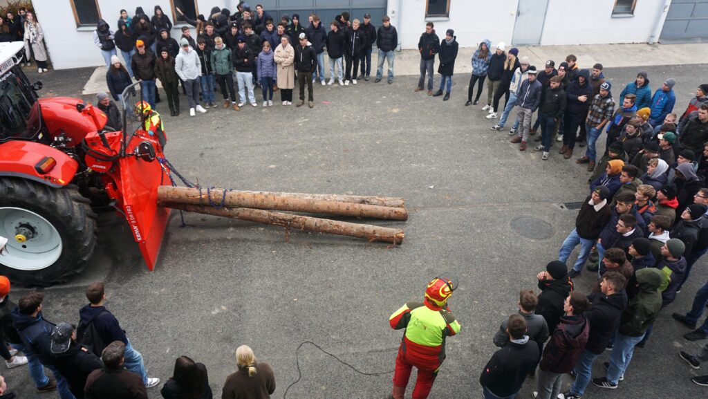 In einem Kreis von zusehenden Schüler:innen befindet sich ein Traktor mit angehängten Baumstämmen udn ein mann in persönlciher SChutzausrüstung.