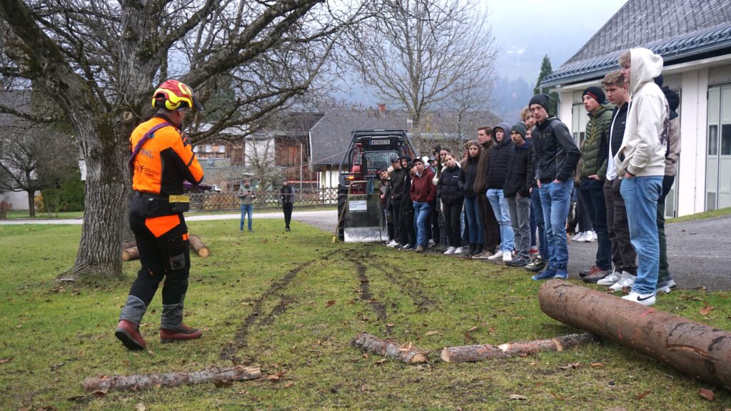 Ein Mann in persönlciher Schutzausrüstung steht vor einer Reihe von Schüler:innen im Freien und zeigt etwas vor.